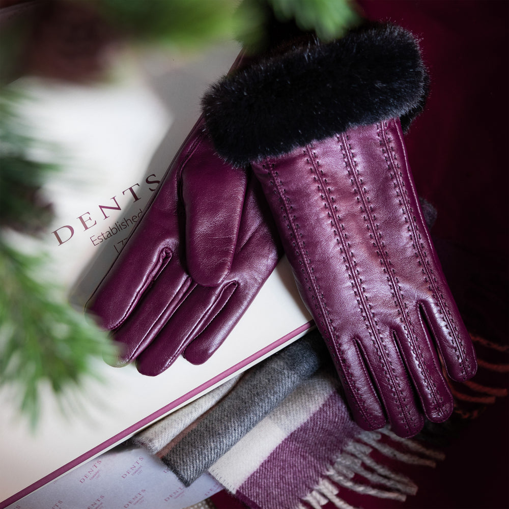 Women's purple leather gloves with black faux fur cuffs and a purple checked cashmere scarf and gift box under a Christmas tree