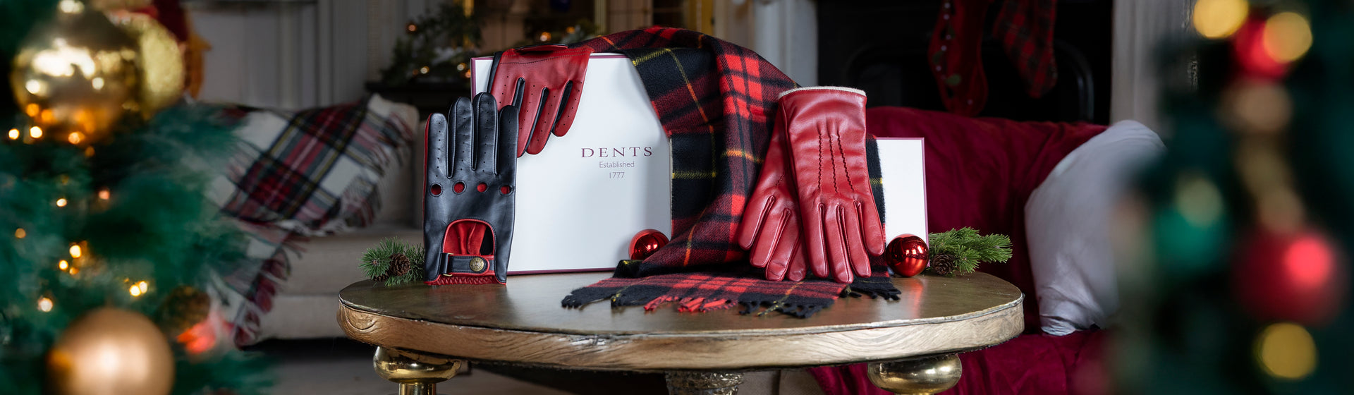 Red women's leather gloves, men's black and red leather driving gloves and a red checked cashmere scarf and gift box on a table with a festive background