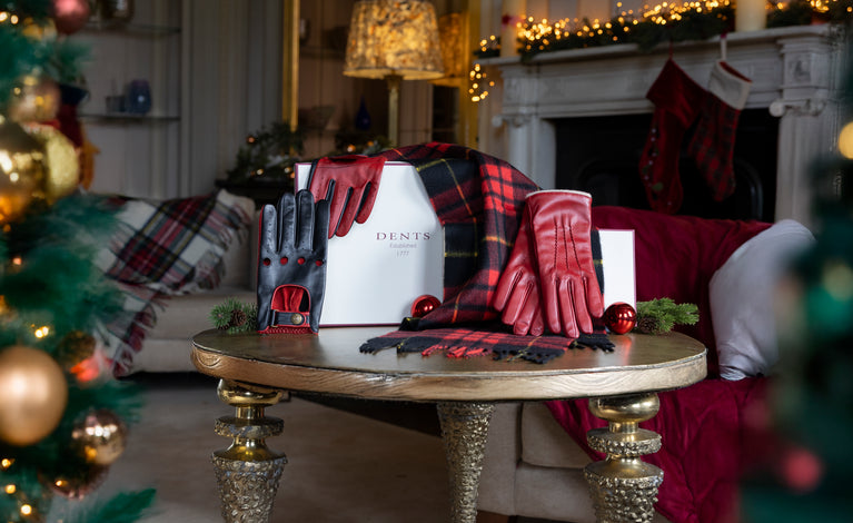 Red women's leather gloves, men's black and red leather driving gloves and a red checked cashmere scarf and gift box on a table with a festive background