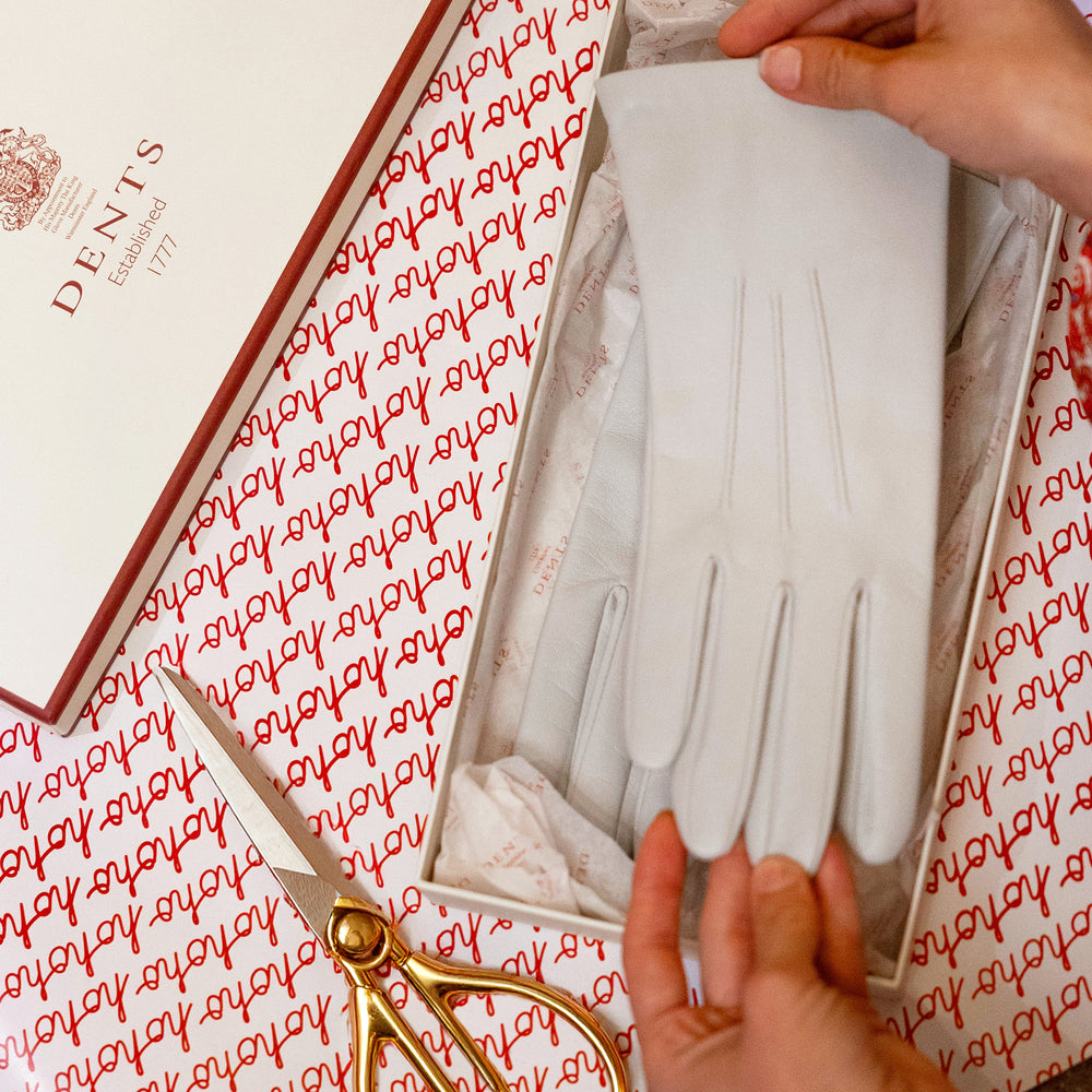 Close-up on hands wrapping a pair of women's white leather gloves into a Dents gift box, with gold scissors and wrapping paper 