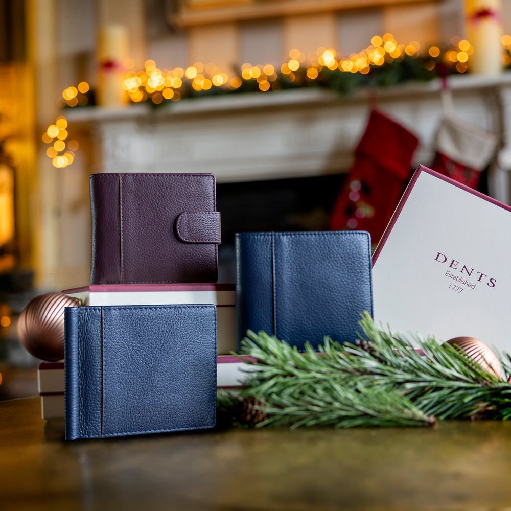 Two men's blue leather wallets and one purple leather wallet on a table with a Dents gift box and green foliage in front of a festive background