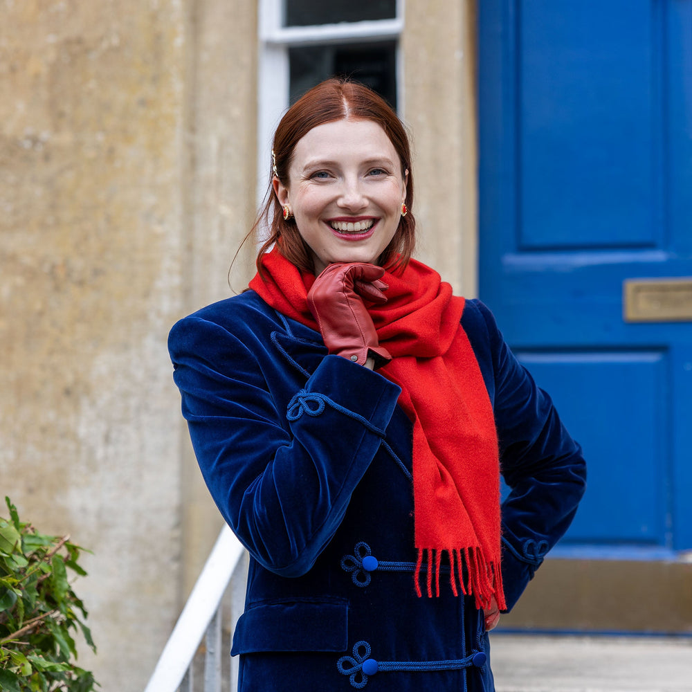 Woman wearing plain cashmere scarf in red in a blue coat standing next to a blue door. 